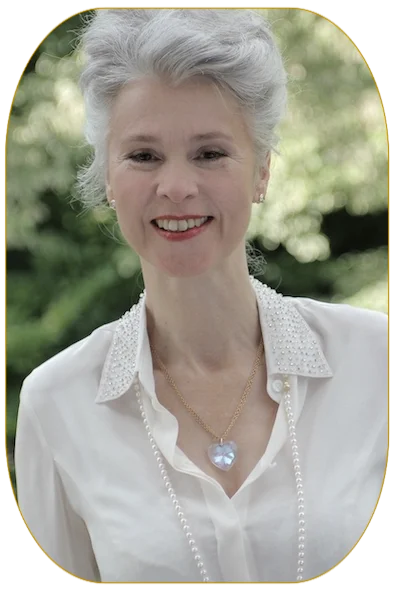 Frau mit hochgesteckten weißen Haaren und  weißer Bluse mit Herzkette und Perlenkette  vor grüner Natur